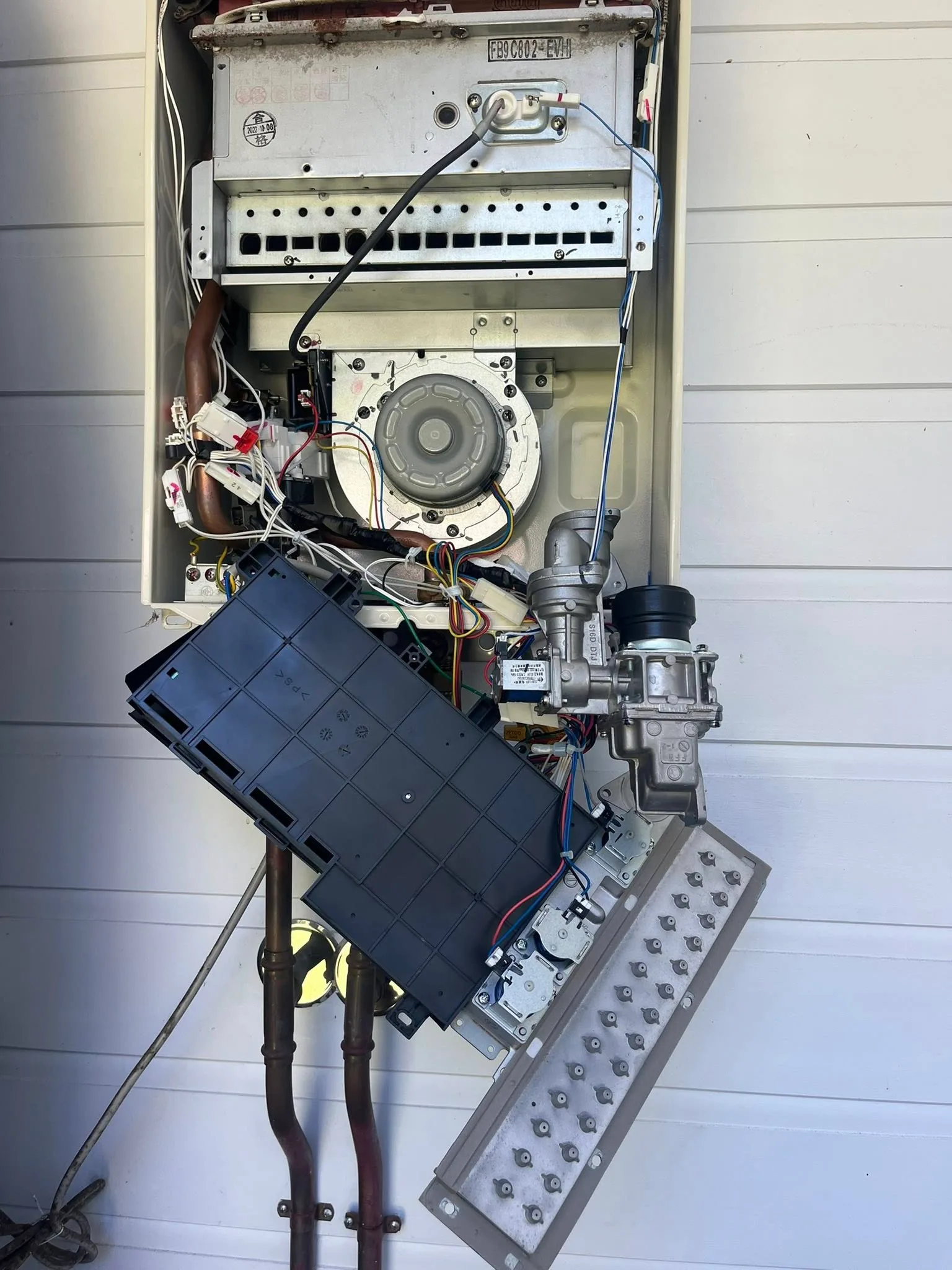 A gas hot water unit on a weatherboard wall with its front cover off and the control box swung out.