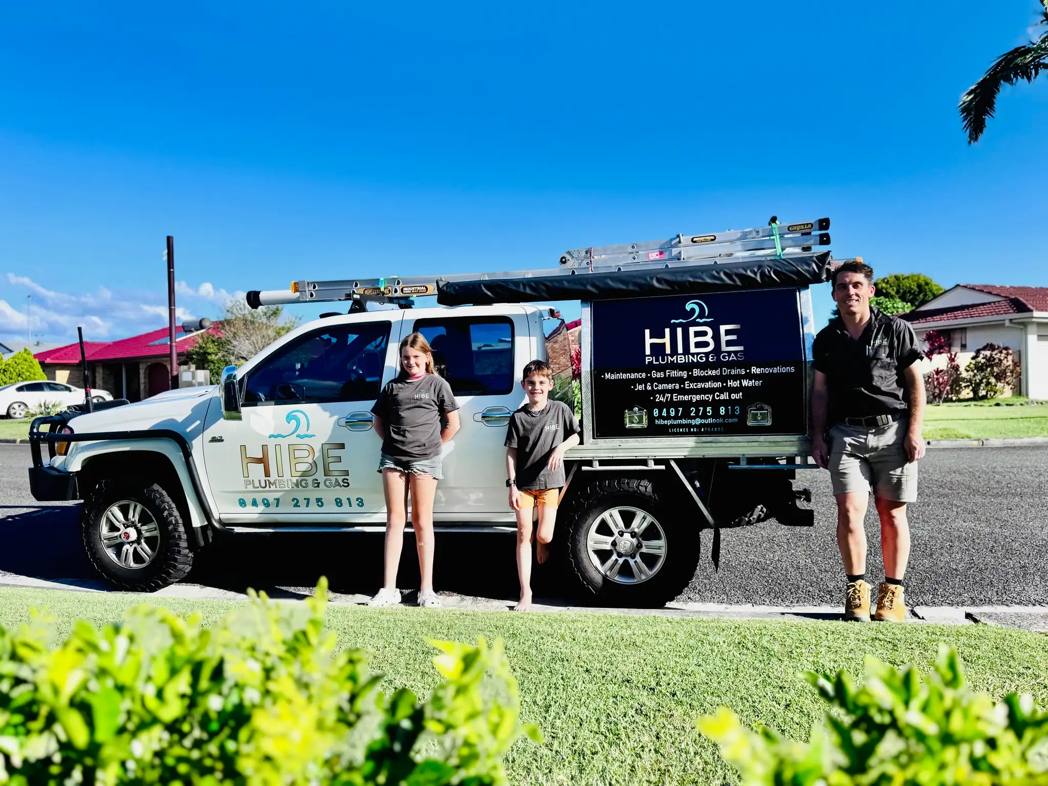 A man and two children standing in front of the white HIBE Plumbing & Gas ute on a suburban street.