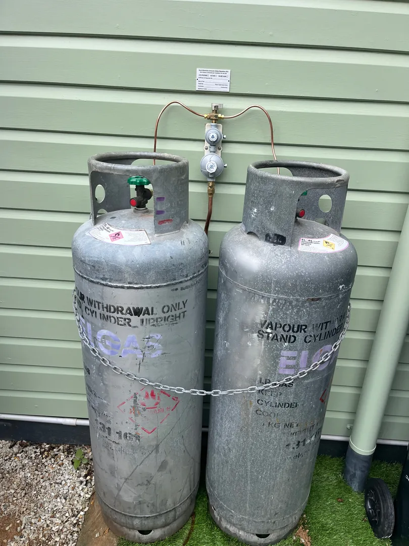 Two grey LPG cylinders chained to a green wall, joined by copper pipes to a regulator between them.